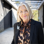 photograph of Tracee smiling, she has short blonde hair and is wearing a black blazer with a cheetah print blouse. She is standing outside, in front of a modern building